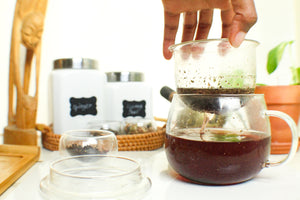 Hand pouring coffee into a glass carafe on a table with jars and a plant in the background.